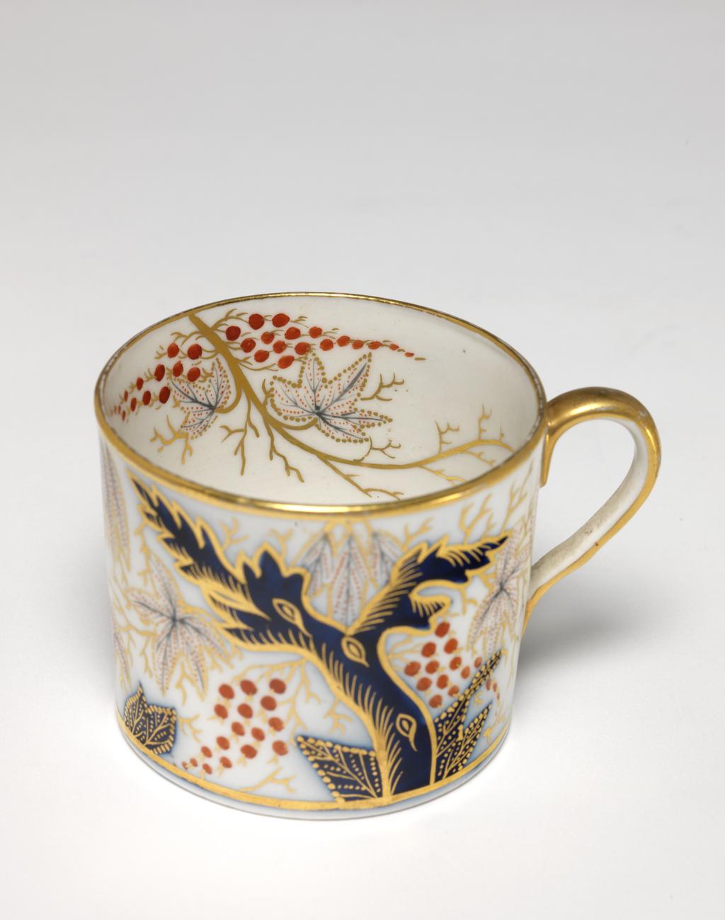 An image of Coffee cup and saucer. New Hall Porcelain Factory, Staffordshire. Cylindrical coffee can with loop handle. Circular saucer with curved sides, standing on a footring. The cup is decorated on the exterior with a blue tree, with foliage dotted in blue and red, and with two bunches of red berries, and gold tendrils, repeated on the other side. Inside there are two gold branches, each with two dotted leaves, and two bunches of red berries. There are gold bands round the rim and base and the back of the handle is gilded. The saucer has a central gold-edged medallion containing a plant, surrounded by the pattern on the exterior of the cup. The edge is encircled by a gold band. Pattern no. 372. Hybrid hard-paste porcelain, painted underglaze in enamels,  decoration in cobalt blue, and gilded, height, cup, 5.9 cm, width, cup, 8.8 cm, diameter, cup, 6.8 cm, height, saucer, 3.1 cm, diameter, saucer, 13.6 cm, circa 1795-1800. Oriental Style.