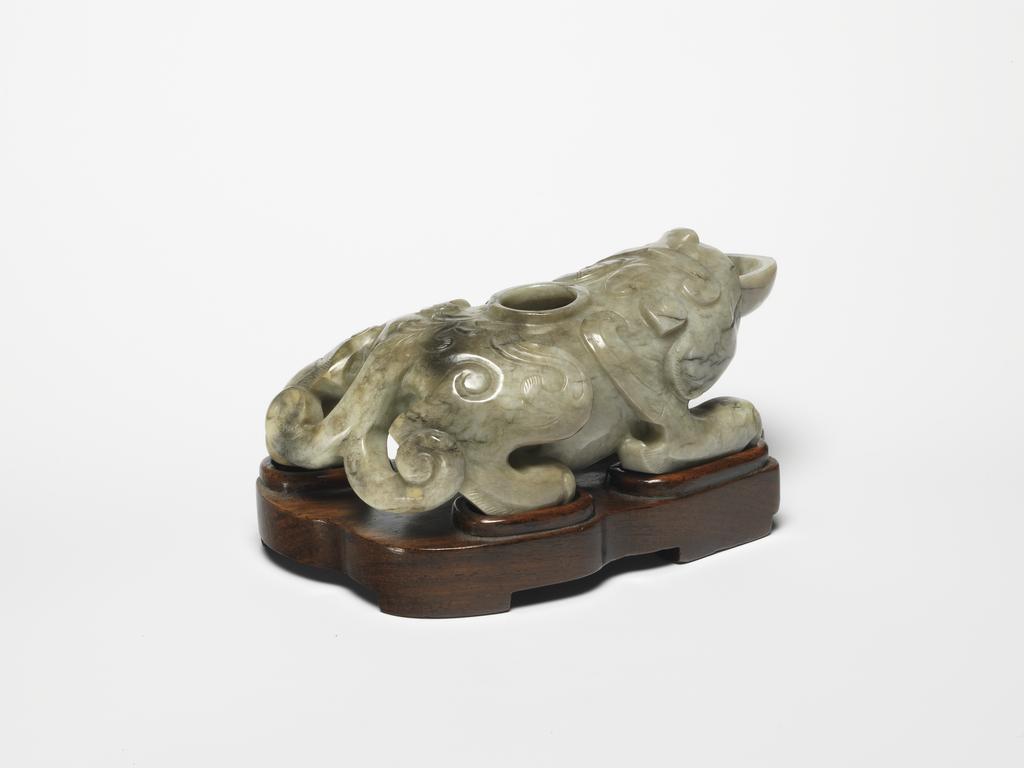 An image of Water Dropper. Unknown carver. Jade with areas of black, carved with mythical beast holding a shallow bowl. Wings extend from the four legs and a hole was drilled in the middle of its back. Jade, greyish, 1500-1600. Chinese. No dimensions recorded.