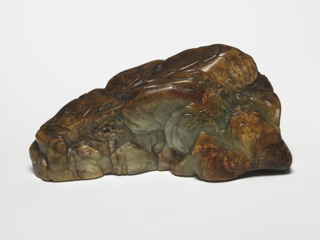 An image of Ornament/Boulder. Front of object. Carved with a Da Mo (Bodhidarma) seated inside a cave among craggy mountains. Green jade with large brown areas, height 7.8 cm, length 16.8 cm, 1700-1800. Chinese. Notes: Da Mo was the founder and first patriarch of Chan/Zen Buddhism. In 527, 32 years after the founding of Shaolin Temple, Bodhidharma arrived in Canton province in China. In China he was referred to as Da Mo. Behind the Shaolin Temple there are five breast-shaped mountains. When the Shaolin monks invited Da Mo to come stay at the temple, Da Mo did not reply. Instead, he immediately went to a cave which was located on one of the breast-shaped mountains. Inside the cave Da Mo sat down facing a wall and immediately began meditating. He meditated for nine years. During the years of meditation, Shin Huang stayed outside Da Mo’s cave and protected Da Mo from any wild animals or other threats. A poem was incised on the rock outside the cave. The inscription for this carving may have been a woodblock print on the theme in the 18th century.
