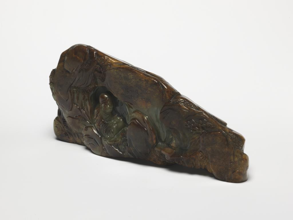 An image of Ornament/Boulder. Front of object. Carved with a Da Mo (Bodhidarma) seated inside a cave among craggy mountains. Green jade with large brown areas, height 7.8 cm, length 16.8 cm, 1700-1800. Chinese. Notes: Da Mo was the founder and first patriarch of Chan/Zen Buddhism. In 527, 32 years after the founding of Shaolin Temple, Bodhidharma arrived in Canton province in China. In China he was referred to as Da Mo. Behind the Shaolin Temple there are five breast-shaped mountains. When the Shaolin monks invited Da Mo to come stay at the temple, Da Mo did not reply. Instead, he immediately went to a cave which was located on one of the breast-shaped mountains. Inside the cave Da Mo sat down facing a wall and immediately began meditating. He meditated for nine years. During the years of meditation, Shin Huang stayed outside Da Mo’s cave and protected Da Mo from any wild animals or other threats. A poem was incised on the rock outside the cave. The inscription for this carving may have been a woodblock print on the theme in the 18th century.