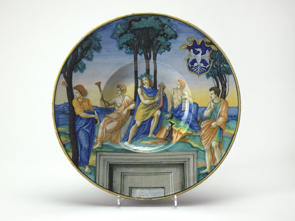 An image of Maiolica dish, painted in polychrome with Mount Parnassus, Apollo, Euterpe and Erato. Milan Marsyas Painter, probably, Italy, The Marches, Urbino. Shape 55. Circular with broad, sloping rim, shallow well and convex centre. Parnassus. In the middle, Apollo sits playing a lyre in front of three trees. On the left, the Muse Euterpe sits holding a trumpet, and on the right, the Muse Erato, holding pipes. On each side a poet stands beside a tree; the one on the left looks away from the central group, the other towards it, holding his right index finger to his lips. Behind the figures there is a landscape and below, a window frame. On the upper right side there is a shield of Tuscan form, charged with the arms azure, an eagle displayed argent. The edge is yellow. Pale yellowish buff earthenware, tin-glazed overall. There is a fault in the glaze on the left of the rim and on the reverse it is pale beige and thinly applied. Painted in blue, green, yellow, orange, brown, black, grey, and white. Height, whole, 4.1 cm, diameter, whole, 30.7 cm, circa 1530-1531. Renaissance. Notes: The scene was derived from the engraving by Marcantonio Raimondi after a preparatory drawing by Raphael for his fresco of Parnassus, in the Stanza della Segnatura in the Vatican. All the figures other than Apollo, two Muses and two poets have been omitted; the trees and, curiously, the window frame, have been retained, and a landscape background has been inserted.