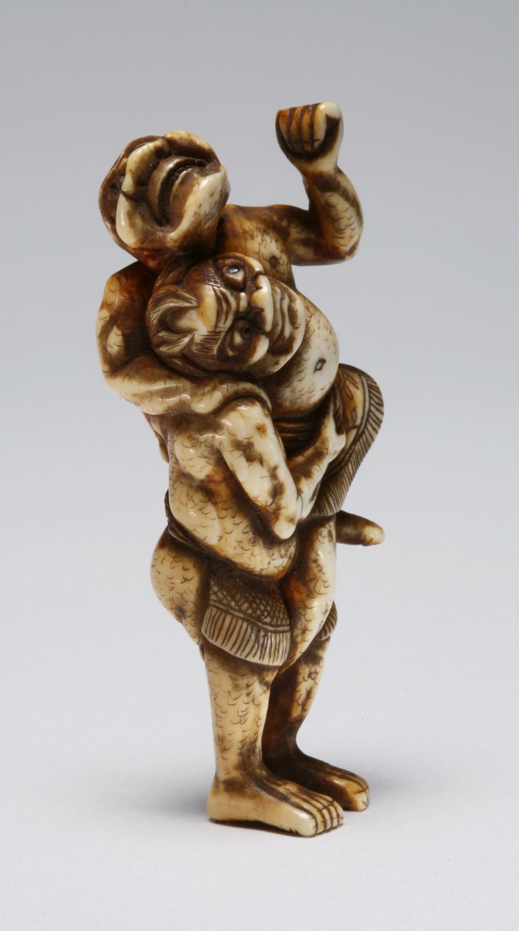 An image of Netsuke. Unknown maker, Japan. Two Sumo wrestlers, one is lifting his opponent up and attempting a throw by grabbing his belt and left leg. Both men’s eyes are inlaid with horn. Ivory, carved, 1800-1850.