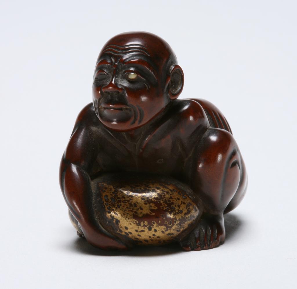 An image of Netsuke. Unknown maker, Japan. A blind man and stone lifter crouching down trying to lift up a large boulder with both hands. Boxwood, ivory and gold lacquer, 1830-1870.