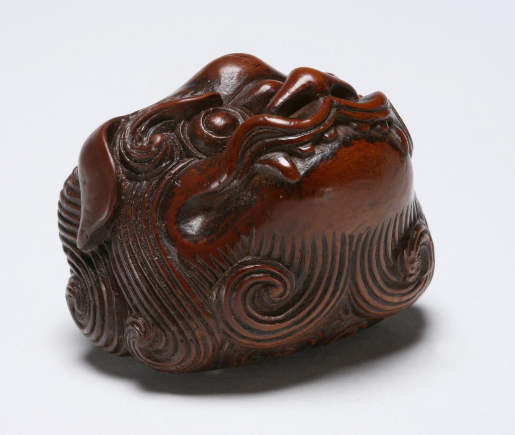 An image of Netsuke. Unknown maker, Japan. A head of Shishi with elaborate curls of hair. Boxwood, caved, circa 1830.