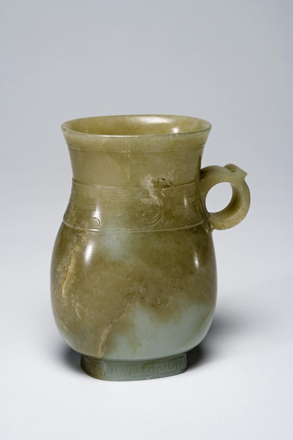 An image of Tankard/Jug. Decorated with a band of archaistic bronze motif around the neck. Pale taupe nephrite with incised decoration. Of oval baluster form with a ring handle and oval footring. Decorated round the rim with a narrow band of key ornament and at the level of the handle with a wider band of circles containing C-scrolls, and areas of hatching. The footring is decorated with key ornament. Height 10.6 cm. Probably Song Dynasty (960-1279). Chinese.