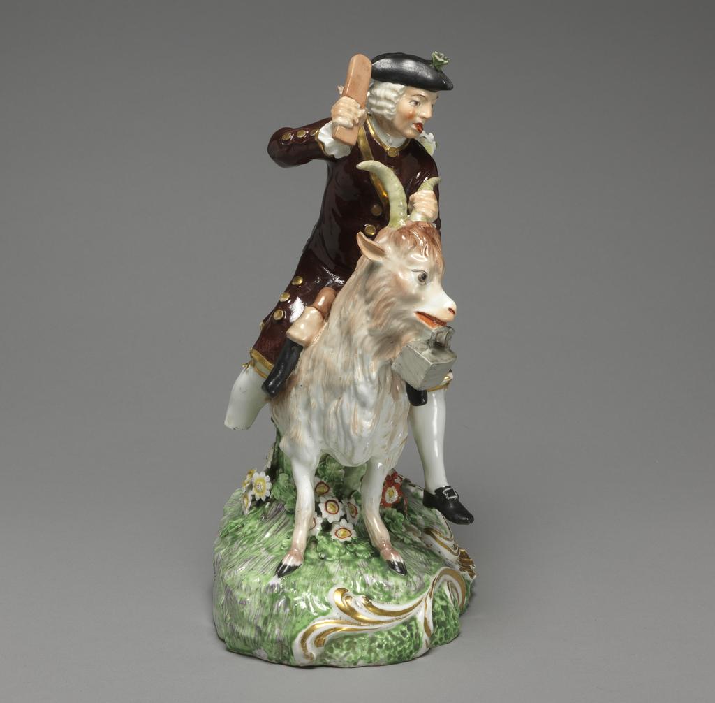 An image of Figure. Taylor riding on a Goat. Derby Porcelain Factory, England, Derbyshire. Kändler, Johann Joachim, modeller, after (German, 1706-1775). Eberlein, Johann Friedrich, modeller, after (German, 1696-1749). The rounded oval mound base is moulded round the front with a rocaille frill flanked by scrolls, and rises up in the middle into a circular green stump decorated with four clumps of applied flowers and leaves. The taylor rides on a Billy goat which is going to the viewer’s left, supported underneath by the stump. He holds the goat’s left horn in his left hand and in his raised right hand holds a bat. He has a ruddy complexion, red lips, and a white wig. He wears a black tricorn hat, a dark brown coat with gold edges and buttons, gold garters at the knee of his beeches, and black shoes with buckles. A basket containing two kids is strapped to his back, and a pair of scissors, two small bags, and some rolled white material protrude from his left pocket (facing the viewer). The goat is painted in shades of brown, and has yellowish-grey horns, and black hooves. A pair of pistols is suspended in holsters on either side of its neck, and it carries a flat iron in its mouth. The base is coloured green and brown, the flowers are white and red with yellow centres, white and blue with yellow centres, white with yellow and red centres, and white with yellow centres. The scrolls and ‘frill’ are picked out in gold. Soft-paste porcelain, slip-cast, with applied hand-modelled details, lead-glazed, and painted overgraze in enamels, and gilt. The underside has a large, circular, central ventilation hole, and has been ground down to level it so that very little glaze remains. Height, whole, 24.2 cm, length, base, 18 cm, circa 1800. Rococo.