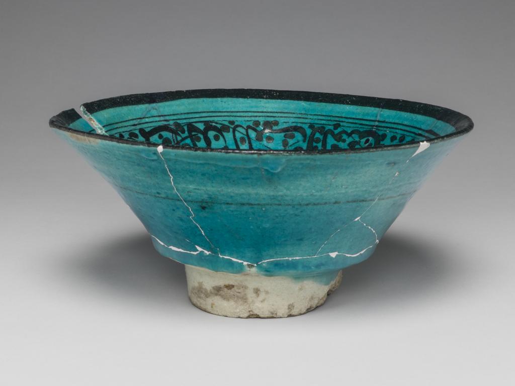 An image of Islamic pottery. Bowl. Unknown potter, Syria, probably Raqqa. Shape: bi-conical bowl on a narrow, high foot ring. Interior: on the rim a concentric band, on the body a band of cursive script is painted on a dotted, crossed background. In the roundel a filler pattern is visible but the remainder obscured by sherds, which have fallen in the bowl from another vessel, during firing. Exterior: at a mid point on the body a thin black line is painted. Glaze runs over the surface, pooling at the carination, the remainder is coated with a thinner glaze. Fritware, wheel thrown, painted in black under a crazed, turquoise glaze, height, whole, 9 cm, diameter, rim, 19.5 cm, diameter, base, 7 cm, weight, whole, 345 g, circa 1200- circa 1250. Ayyubid.