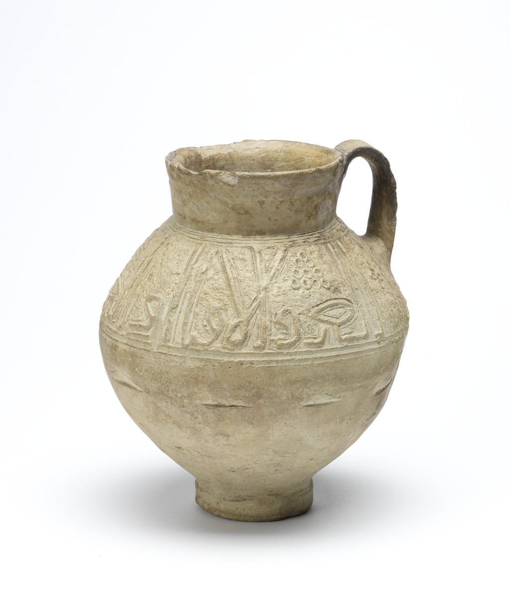 An image of Islamic pottery. Jug. Unknown potter, probably, Iran or Syria. Shape: oval body on a disc base with a short, slightly flaring neck and a strap handle attached to the neck and shoulder. Exterior: a frieze of kufic script is moulded on the shoulder below which a series of impressed marks indicates the process of joining this decoration to the body of the jug. On the neck an impressed concentric band is visible. Interior: undecorated. Buff marl earthenware, probably wheel thrown with moulded decoration applied on the shoulder. Height, whole, 16.8 cm, width, whole, 14.7 cm, diameter, rim, 8.2 cm, diameter, base, 5.5 cm, weight, whole, 652 g, circa 1100-circa 1299. Ayyubid, Seljuk.