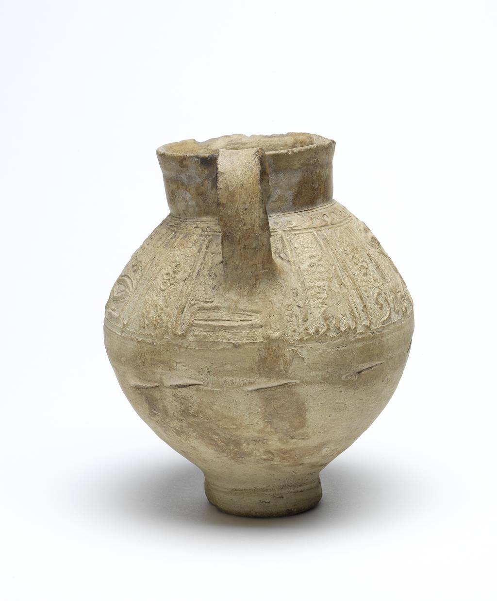 An image of Islamic pottery. Jug. Unknown potter, probably, Iran or Syria. Shape: oval body on a disc base with a short, slightly flaring neck and a strap handle attached to the neck and shoulder. Exterior: a frieze of kufic script is moulded on the shoulder below which a series of impressed marks indicates the process of joining this decoration to the body of the jug. On the neck an impressed concentric band is visible. Interior: undecorated. Buff marl earthenware, probably wheel thrown with moulded decoration applied on the shoulder. Height, whole, 16.8 cm, width, whole, 14.7 cm, diameter, rim, 8.2 cm, diameter, base, 5.5 cm, weight, whole, 652 g, circa 1100-circa 1299. Ayyubid, Seljuk.