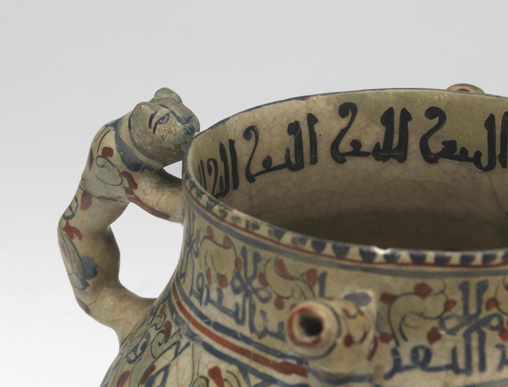 An image of Islamic pottery, Minai. Posset-pot. Unknown potter, Iran, central Asia, probably Kashan. Shape: bulbous body with a cylindrical neck and plain rim, sits on a low foot ring. Opposing cheetah handles and angular spouts are attached to the upper body. Exterior: on the rim a dentilated pattern is painted in blue. On the neck an epigraphic band is painted in blue and is interspersed with turquoise leaves and red dots. On the body arabesques of turquoise half-palmettes, sketchy blue birds and red dots covers the surface. On the cheetah handles and the spouts continuation of the patterns from the body are present, except on the head of the animals, which are painted turquoise with black lines to indicate features. Glaze covers the surface to the foot ring where it terminates unevenly. Interior: on the neck an epigraphic band is painted in black. The surface is coated in a glaze but otherwise is undecorated. Fritware, wheel thrown and mould-made, coated in white glaze painted with red, blue, turquoise and black. Height, whole, 14.6 cm, width, whole, 20.7 cm, diameter, rim, 11.4 cm, diameter, base, 8.7 cm, weight, whole, 694 g, circa 1170- circa 1220. Seljuk.