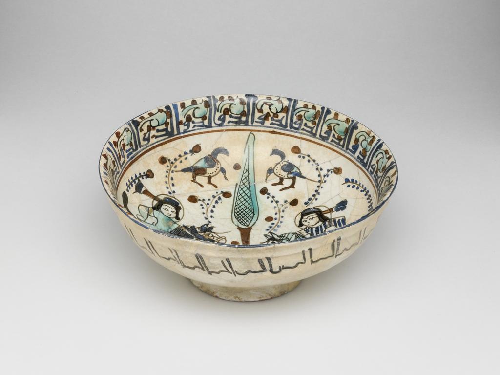 An image of Bowl. Islamic pottery. Minai. Unknown, potter, central Asia, probably Kashan. Shape: carinated bowl with collared neck, sits on a slightly flaring foot ring base. Interior: on the neck a kufic inscription is painted in blue and surrounded by turquoise leaves and red dots. On the body two mounted horsemen, wearing headdresses and tunics with gilding, are confronted either side of a cypress tree surrounded by leafy fronds dotes with blue and red. Above and below the horsemen pairs polychrome birds are painted. Exterior: on the upper body a cursive inscription is painted in black. Glaze covers the surface unevenly, terminating unevenly on the foot ring. Fritware, wheel thrown, coated in white glaze painted with dark red, blue, turquoise, black and gilded with gold leaf,. Height, whole, 7.4 cm, width, whole, 16.8 cm, diameter, rim, 16.8 cm, diameter, base, 7.2 cm, weight, whole, 242 g, circa 1170- circa 1220. Seljuk Period.