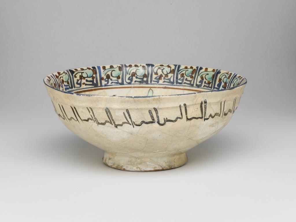 An image of Bowl. Islamic pottery. Minai. Unknown, potter, central Asia, probably Kashan. Shape: carinated bowl with collared neck, sits on a slightly flaring foot ring base. Interior: on the neck a kufic inscription is painted in blue and surrounded by turquoise leaves and red dots. On the body two mounted horsemen, wearing headdresses and tunics with gilding, are confronted either side of a cypress tree surrounded by leafy fronds dotes with blue and red. Above and below the horsemen pairs polychrome birds are painted. Exterior: on the upper body a cursive inscription is painted in black. Glaze covers the surface unevenly, terminating unevenly on the foot ring. Fritware, wheel thrown, coated in white glaze painted with dark red, blue, turquoise, black and gilded with gold leaf,. Height, whole, 7.4 cm, width, whole, 16.8 cm, diameter, rim, 16.8 cm, diameter, base, 7.2 cm, weight, whole, 242 g, circa 1170- circa 1220. Seljuk Period.