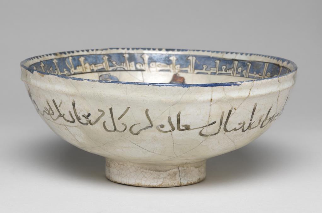 An image of Bowl. Islamic pottery, Minai. Unknown potter, central Asia, probably Kashan. Shape: hemispherical bowl with collared neck, sits on a low foot ring. Interior: the rim is painted with a blue dentilated pattern. On the neck and around the edge of the roundel, a kufic inscription is outlined in black and reserved in blue. On the body pairs of opposed, gesticulating male figures wear blue, purple or green patterned robes with gilding and red, purple or blue hats. Green tear-drop cartouche adorned with blue or red circles separate the pairs of figures. On the base, a mounted horseman, dressed in purple robes, rides a blue dappled horse and supports two hawks. Exterior: the rim is painted blue, on the neck a naskh inscription is painted in black. Glaze covers the exterior surface evenly except the underside of the foot ring. Fritware, wheel thrown, coated in a white glaze painted in six colours and gilded. Height, whole, 8 cm, diameter, rim, 18.8 cm, diameter, base, 6.8 cm, weight 336 g, circa 1170-circa 1220. Seljuk.