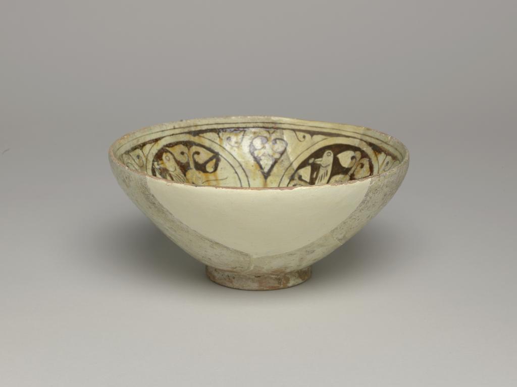 An image of Bowl. Islamic pottery. Garrus ware. Unknown potter, Iran. Shape: hemispherical bowl with a plain rim on a disc base cut, away on the underside. Interior: on the body a frieze of four circular medallions, separated by palmettes, contains birds and scrolling leaves, all carved through a cream slip. The glaze pools and is cracked in the base, splashes of green pigment are present in one area. Three scar marks on the lower body indicated the use of a tripod stacking device in the kiln. Pinkish earthenware, wheel thrown, coated in a white slip carved under a colourless glaze, splashed with traces of green. Exterior: undecorated except for a cream slip and colourless glaze extending to the foot ring. Height, whole, 8.5 cm, width, whole, 18.6 cm, diameter, base, 6.8 cm, circa 1100-circa 1299. Seljuk.