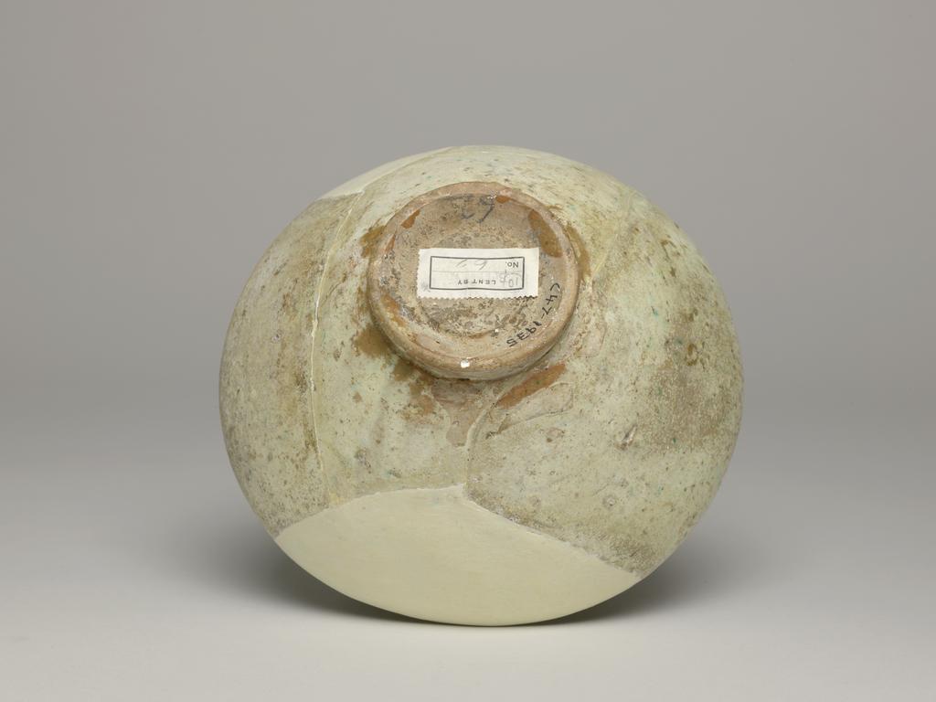 An image of Bowl. Islamic pottery. Garrus ware. Unknown potter, Iran. Shape: hemispherical bowl with a plain rim on a disc base cut, away on the underside. Interior: on the body a frieze of four circular medallions, separated by palmettes, contains birds and scrolling leaves, all carved through a cream slip. The glaze pools and is cracked in the base, splashes of green pigment are present in one area. Three scar marks on the lower body indicated the use of a tripod stacking device in the kiln. Pinkish earthenware, wheel thrown, coated in a white slip carved under a colourless glaze, splashed with traces of green. Exterior: undecorated except for a cream slip and colourless glaze extending to the foot ring. Height, whole, 8.5 cm, width, whole, 18.6 cm, diameter, base, 6.8 cm, circa 1100-circa 1299. Seljuk.