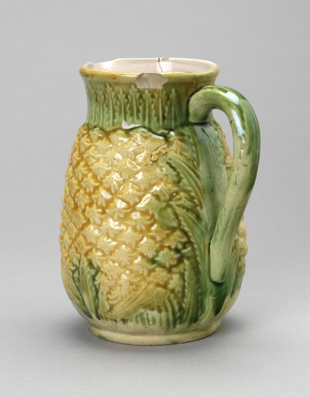 An image of Pineapple jug. Unidentified factory, England. Cream earthenware jug, press moulded in the shape of a pineapple, with a short slightly flaring cylindrical neck, a loop handle and a slim circular foot-rim. The outside is moulded to represent both the fruit and the leaves, the former covered by bright yellow-gold glaze and the latter with a thin green glaze. The interior is tinged pink. Earthenware, press moulded and coated with clear and coloured glazes, height, whole, 14 cm, width, whole, 12 cm, 19th century.