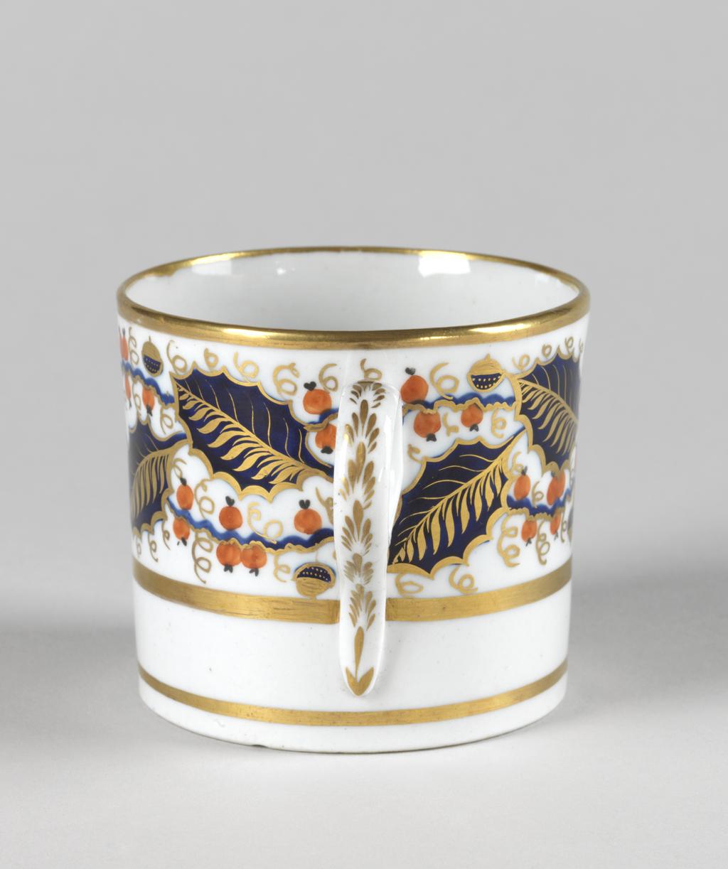 An image of Coffee can. New Hall Porcelain Factory, Staffordshire, New Hall. Painted with a wide band of holly leaves and berries between gold bands. Pattern 927. Cylindrical with a ring handle and slightly recessed base. The upper two-thirds of the exterior is decorated with a broad band of blue and gold holly leaves interspersed with red berries and occasional blue and gold berries on wavy blue stems. There is a broad gold band below, another round the rim, and another just above the lower edge. The back of the handle is decorated in gold with six stylized leaves. Hard-paste porcelain painted in blue under presumed lead-glaze, and in red in overglaze enamel, and gold, height, whole, 6.3 cm, width, whole, 9.2 cm, diameter, whole, 6.9, cm, circa 1800-1810.