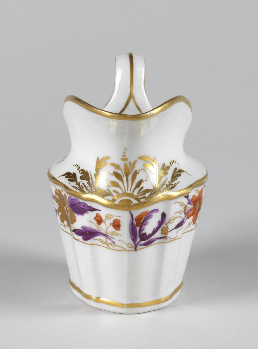 An image of Milk jug. New Hall Porcelain Factory, Staffordshire. Pattern no. 317. Of silver-shape with sloping panelled sides, an upward curving lip rising from the shoulder, and high loop handle. Below the shoulder there is a wide border of gold and red flowers with purple leaves with a narrow horizontal band below and a wider band above. On each side of the shoulder there is a gold sprig, and below the lip an arrangement of stylized gold foliage. A gold line runs around the upper edge and down both sides of the handle and another encircles the inside of the upper edge, then joins at the back to run down the centre of the handle. Another gold band encircles the body a little way up from the lower edge. The glaze on the base of the interior is speckled with grey. Hybrid hard-paste porcelain moulded, painted overglaze in red and purple enamels, and gilded. Height, whole, 11 cm, length, whole, 11.6 cm, length, base, 6 cm, circa 1790-1800. Neoclassical.