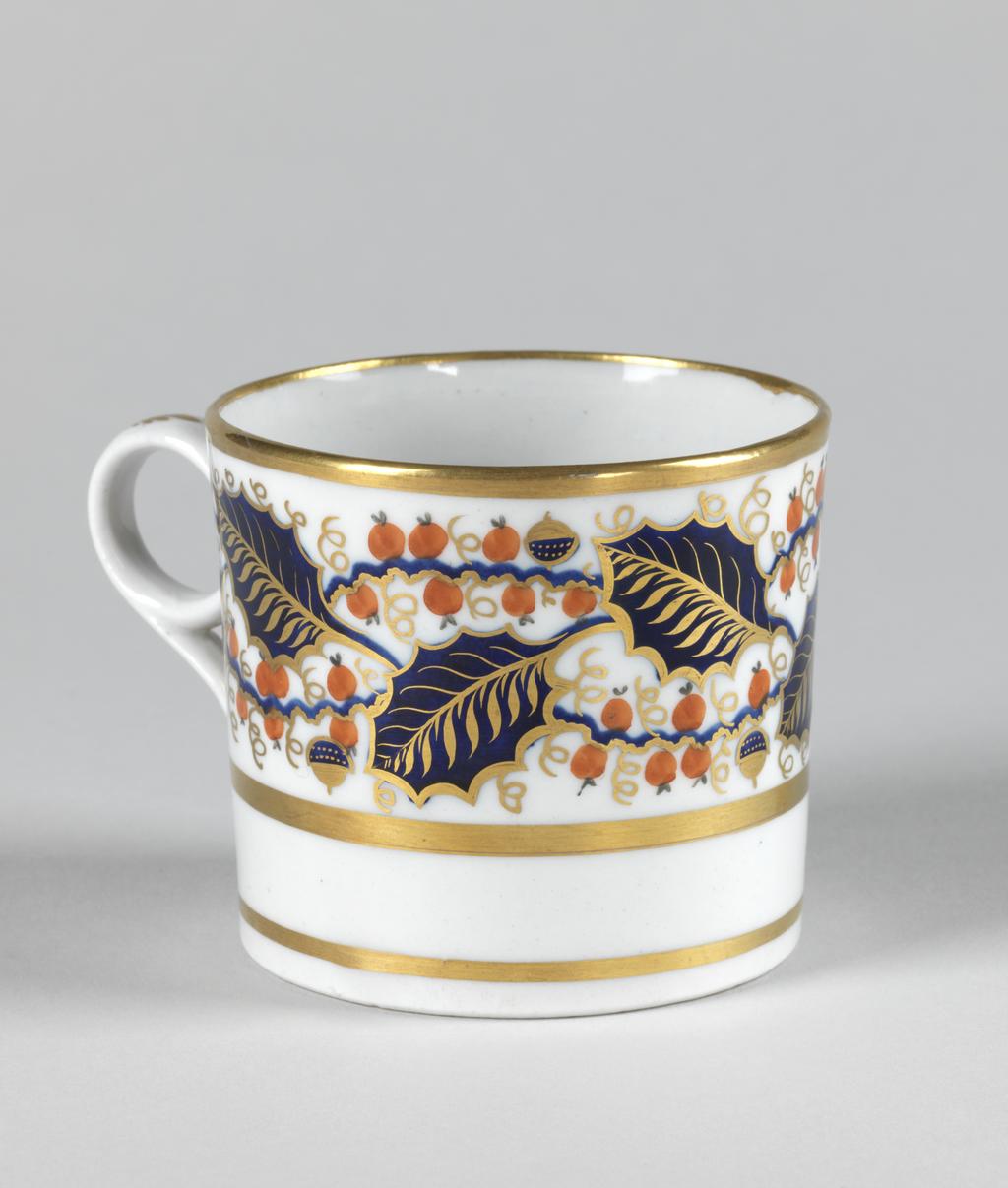 An image of Coffee can. New Hall Porcelain Factory, Staffordshire, New Hall. Painted with a wide band of holly leaves and berries between gold bands. Pattern 927. Cylindrical with a ring handle and slightly recessed base. The upper two-thirds of the exterior is decorated with a broad band of blue and gold holly leaves interspersed with red berries and occasional blue and gold berries on wavy blue stems. There is a broad gold band below, another round the rim, and another just above the lower edge. The back of the handle is decorated in gold with six stylized leaves. Hard-paste porcelain painted in blue under presumed lead-glaze, and in red in overglaze enamel, and gold, height, whole, 6.3 cm, width, whole, 9.2 cm, diameter, whole, 6.9, cm, circa 1800-1810.