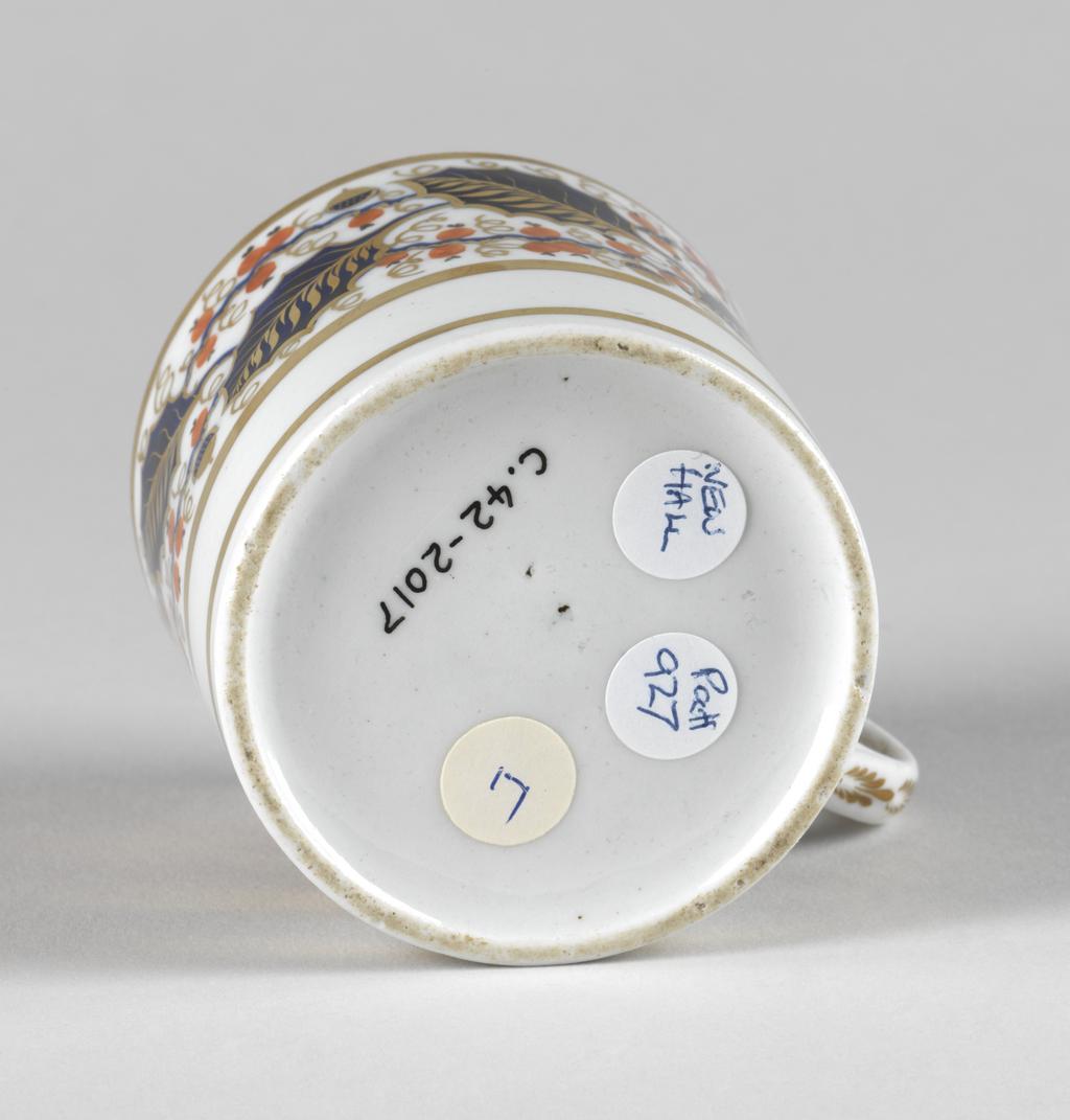 An image of Coffee can. New Hall Porcelain Factory, Staffordshire, New Hall. Painted with a wide band of holly leaves and berries between gold bands. Pattern 927. Cylindrical with a ring handle and slightly recessed base. The upper two-thirds of the exterior is decorated with a broad band of blue and gold holly leaves interspersed with red berries and occasional blue and gold berries on wavy blue stems. There is a broad gold band below, another round the rim, and another just above the lower edge. The back of the handle is decorated in gold with six stylized leaves. Hard-paste porcelain painted in blue under presumed lead-glaze, and in red in overglaze enamel, and gold, height, whole, 6.3 cm, width, whole, 9.2 cm, diameter, whole, 6.9, cm, circa 1800-1810.