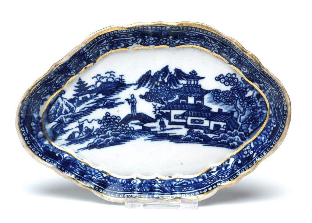 An image of Spoon tray. New Hall Porcelain Factory, England, Staffordshire. Oval with a six-sided shaped edge, shallow, sloping, fluted sides, and flat base. The flat area is decorated with a Chinese landscape, with buildings amid trees on the right, a man near a bridge, and a man standing on a bridge leading to a small island to the left of centre, a boat on water, on the left, a shore with a building amid tree and rocks on the left. On the slides there is border of scrolls, foliage and fruit with dotted beads on the edge. There is a gold band round the lower edge of the sides and round the upper edge. Hybrid hard-paste porcelain printed underglaze in blue, and gilded. Height, whole, 1.4 cm, length, whole, 14.5 cm, width, whole, 9.5 cm, circa 1782-1787. Chinese style.