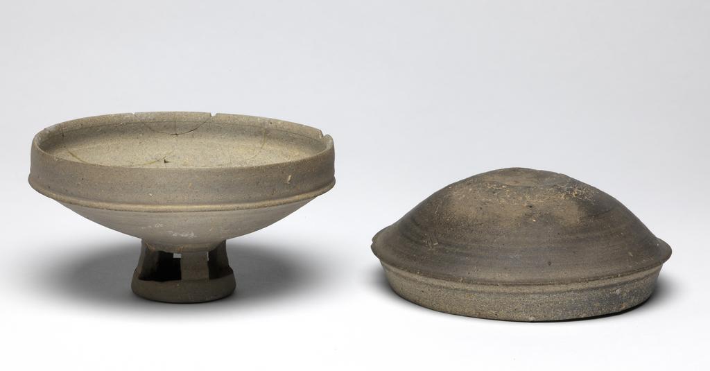 An image of Bowl, on pierced foot with a cover. The bowl and its cover are wide, the stand has four openings. The body is thick and the clay dark grey. Stoneware.. Silla Kingdom, 5th or 6th century.