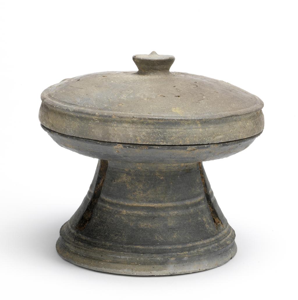 An image of Bowl, on high foot with a cover. Thick and dark grey body. This shallow bowl rests on a tall stem and is divided by horizontal ridges with three long openings. The cover has a mushroom stem knob. Stoneware. Silla Kingdom, 5th or 6th century.