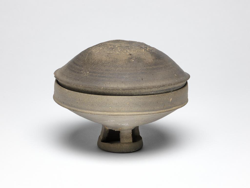 An image of Bowl, on pierced foot with a cover. The bowl and its cover are wide, the stand has four openings. The body is thick and the clay dark grey. Stoneware.. Silla Kingdom, 5th or 6th century.