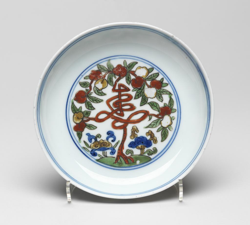 An image of Dish. Production Place: China. A wucai saucer dish, peach tree as Shou (longevity) character (flaking to yellow enamels, slight warp), diameter 14.8 cm. Jiajing period (1522-1566).