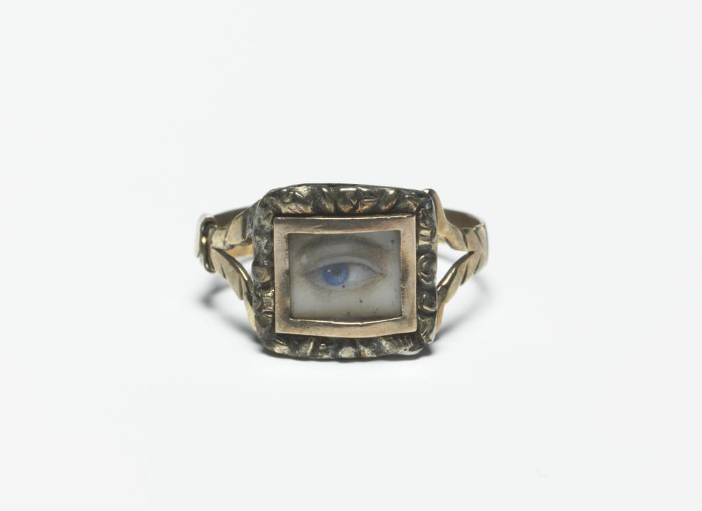 An image of Jewellery/Ring. The ring has a thin gold hoop dividing into two branches at the shoulders, and a rectangular silver bezel with cast and chased border, in which is set a rectangular, plain gold frame enclosing under glass a miniature of a brown eyebrow and an eye with a blue iris. Gold, silver, glass and painted miniature on ivory, height, whole, 1.9 cm, circa 1790-1800. English.