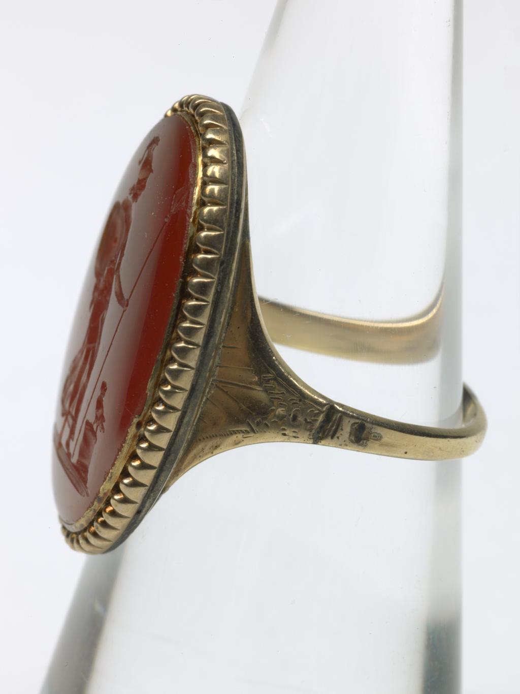 An image of Jewellery. Ring. Intaglio seal. Unknown maker, probably English. Red carnelian intaglio depicting standing figure of Minerva, wearing plumed helmet and chiton, leaning on a spear and carrying her shield on her arm, head turned towards the left, owl on a low altar beside her. Pearled border and ground line. The device also appears on the reverse of a medallic portrait of Gustavus III of Sweden. Gold with pointed oval bezel, beaded border and closed back; square hoop expanding at the shoulders, ornamented with bright cutting. Gold ring, red carnelian intaglio, height, intaglio, 22 mm, width, intaglio, 14 mm, circa 1700-1800.
