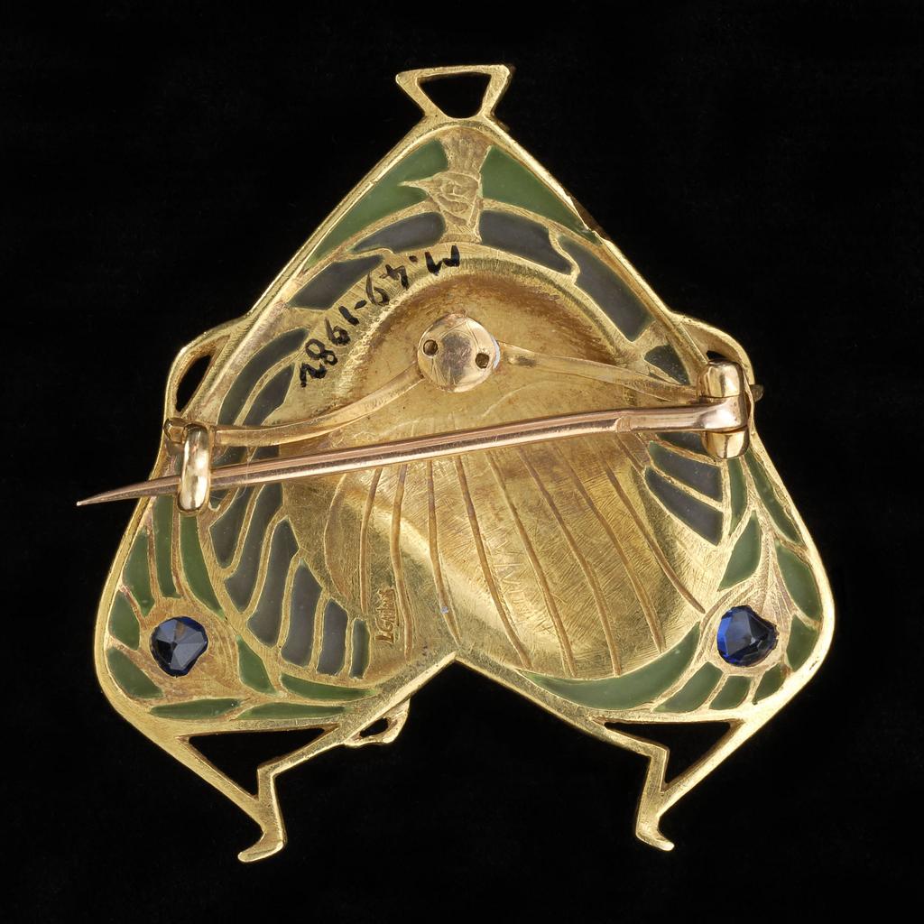 An image of Jewellery/brooch/pendant. Gautrait, Léopold Albert Marin, maker (French, 1865-1937, Paris, Colmar). Three sided with a triangular loop for suspension at the top, and two 'legs' on the lower edge; detachable pin-fastening on the back. In the centre is the head of a woman in profile to right earing a peacock headdress, the tail feathers of which fan out to fill the outline, the sapphires forming the 'eyes'. On the back, engraved lines fill the peacock's outline. Has unaccessioned case not original. Gold frame, green, blue and white enamel, and green and grey translucent plique-à-jour enamel, set with two sapphires. Height, whole, 4 cm, width, whole, 3.9 cm, circa 1900. Art Nouveau.
