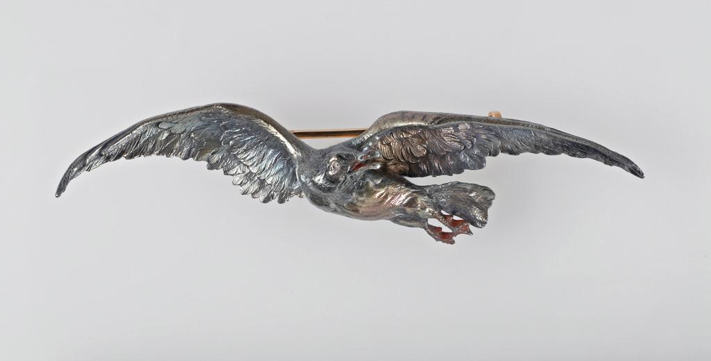 An image of Jewellery. Gull Brooch. Maison Mellerio, Paris, Colmar. Remnants of black enamel on the head and wings, and red enamel on the legs and feet, and inside the beak. Detachable gold pin fastening, inscribed 'MELLERIO.R.PAIX.9.'. Silver, cast and chased, the eye set with a diamond chip, and originally enamelled in black on the head, and red on the legs, feet, and inside of beak. Width, whole, 7.6 cm, circa 1880-1900. Acquisition Credit: Given by Mrs J. Hull Grundy.