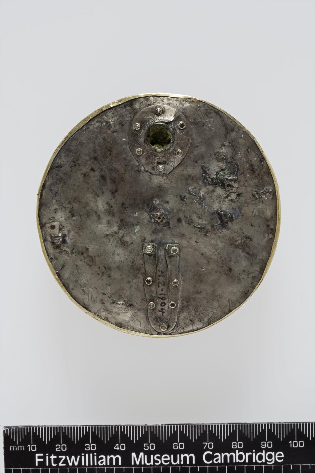 An image of Jewellery/brooch. The front is made of gold plates and the disc at the back of silver. Front: in the centre is a high boss. Round it are four equidistant sockets and between these a cruciform design of garnets on concentric circles. Back: round the edge a band of zoomorphic pattern, similar to that on the back of the Crundale Buckle in the British Musem. The hinge and socket of pin extant. Diameter 8.2cm, thickness 1.1cm, circa 540-560. Anglo-Saxon. Excavated in the 19th century from a cemetery at the King's Field, Faversham, Kent.