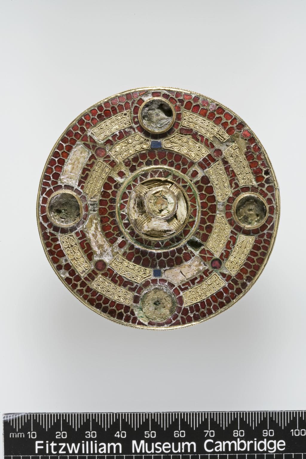 An image of Jewellery/brooch. The front is made of gold plates and the disc at the back of silver. Front: in the centre is a high boss. Round it are four equidistant sockets and between these a cruciform design of garnets on concentric circles. Back: round the edge a band of zoomorphic pattern, similar to that on the back of the Crundale Buckle in the British Musem. The hinge and socket of pin extant. Diameter 8.2cm, thickness 1.1cm, circa 540-560. Anglo-Saxon. Excavated in the 19th century from a cemetery at the King's Field, Faversham, Kent.