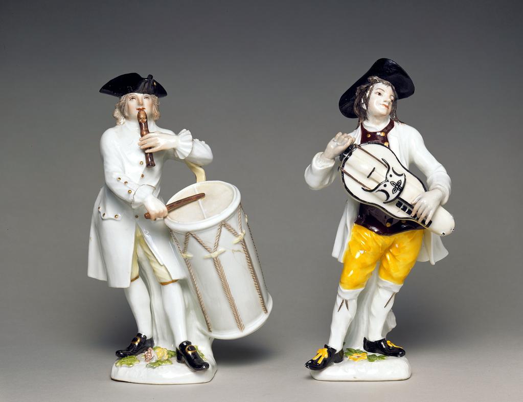 An image of C.32-1954. Provençal Drummer from the Cries de Paris. Meissen Porcelain Factory, Germany. Reinicke, Peter, modeller. The underside of the base is unglazed, and has had the ventilation hole and two firing cracks filled with a brown substance.  The low, irregular four-sided base has a tall tree stump at the back and is decorated with applied leaves and a yellow and a red and white rose at the front. The musician stands on his right leg with his left extended in front. In his left hand he holds a large brown pipe to his lips. In his right hand he holds a brown drum-stick with which he plays on a monstrous white drum decorated with pale brown rope trimmings. The base of the drum rests on the tree-stump, and it is supported by a beige strap which hangs over his left arm. The drummer has thick curly grey hair hanging down to his shoulders. He wears a black three-cornered hat with a gold stud on its left side, a white shirt with ruffles at the wrists, a long white coat with gold buttons; pale yellow breeches with gold bands at the knees; white stockings, and black shoes with gold buckles.  Hard-paste porcelain, press-moulded, and painted overglaze in green, yellow, flesh pink, a little red, beige, brown, and black enamels, and gilt, height 19.5 cm width 12.2 cm, depth 6.5 cm, 1747. Rococo.C.27-1954: Hurdy-gurdy player. Meissen Porcelain Factory, Germany. Reinicke, Peter, modeller. The unglazed base has a small round ventilation hole near the back. The rounded square low mound base has a tree stump at the back, and is decorated on top with applied leaves, and a red, a yellow and a mauve flower. The hurdy-gurdy player stands on his left foot with right leg forward. He carries a large white hurdy-gurdy outlined in black, and with his right hand turns the handle of the wheel which sounds the strings, while with his left hand he plays upon the keys which stop the strings and thus obtain the notes. He has long brownish-black hair trailing untidily down his back, and wears a b